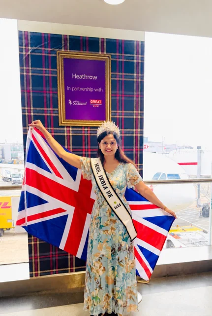 Dr Sivasankari Babu holding a Union flag at Heathrow Airport, wearing a crown and a sash reading Mrs India UK 2026