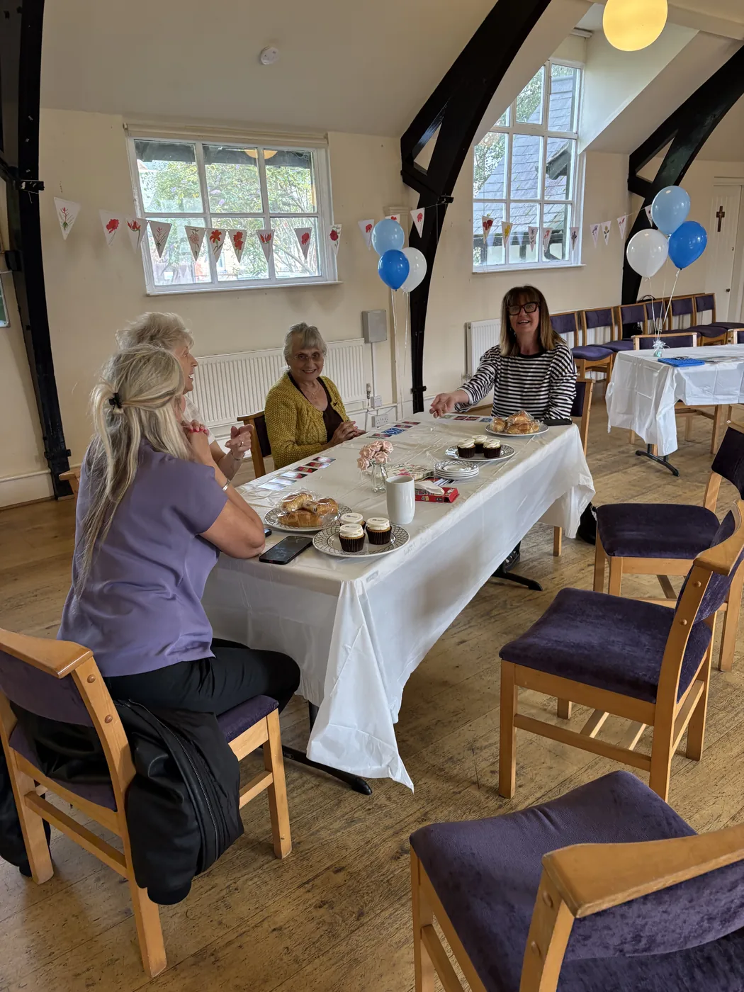 Four people sitting around a long table laid with a white cloth, cakes and scones, in a church hall hung with bunting