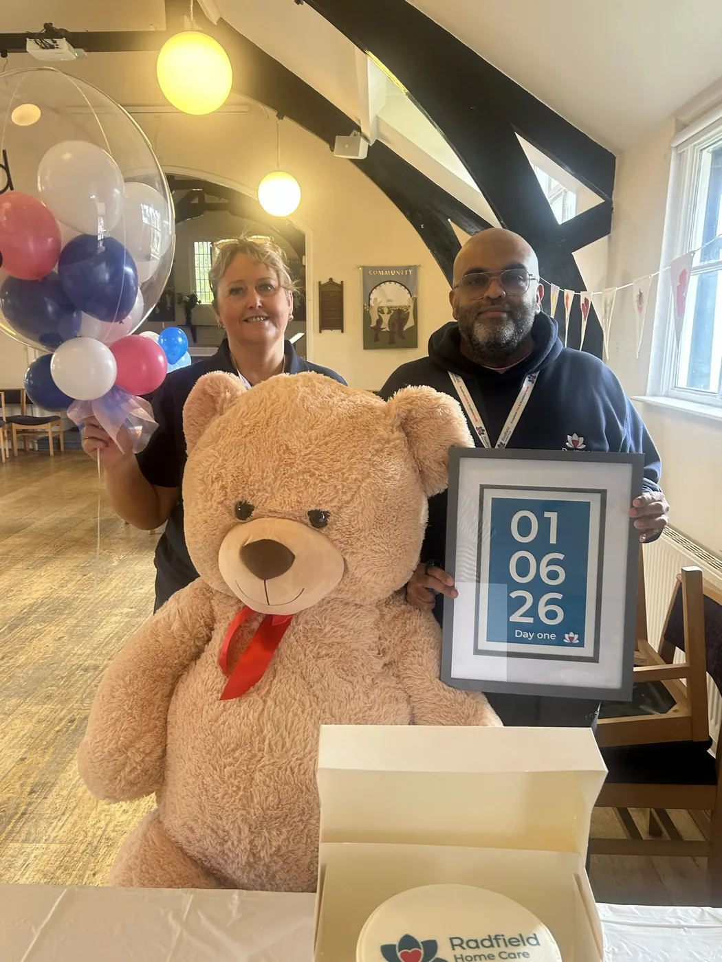 Two people in the church hall holding a large teddy bear and a framed print reading 01 06 26, Day one, beside a Radfield Home Care box
