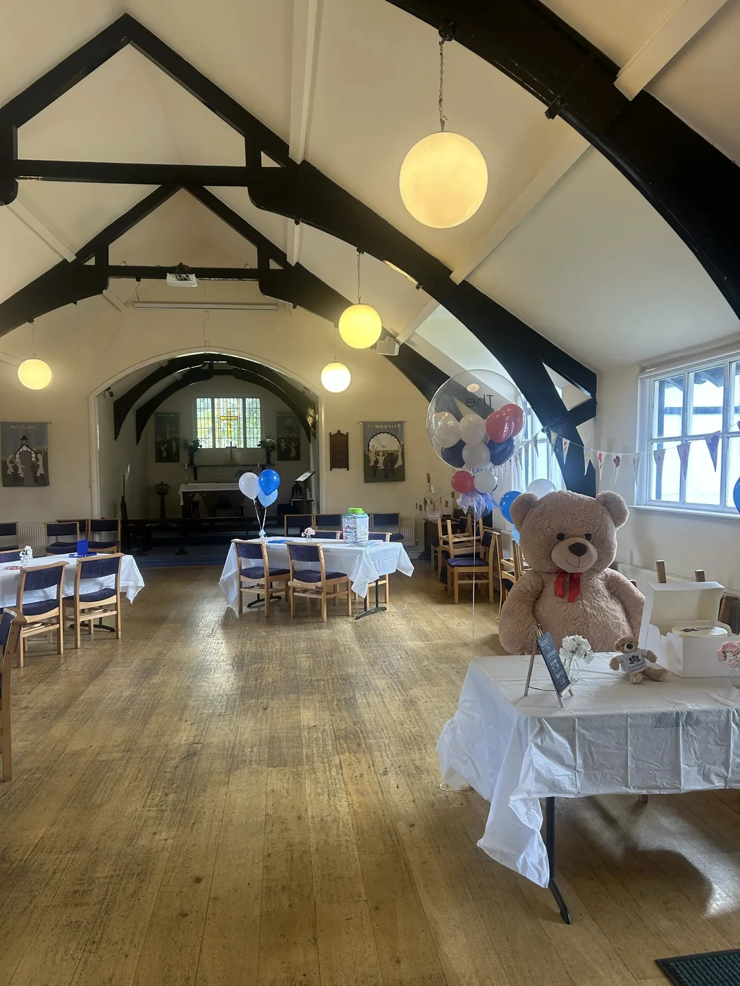 The hall at St Luke's Church, Bowdon, set out with tables under white cloths, balloons and bunting, with a timber roof overhead