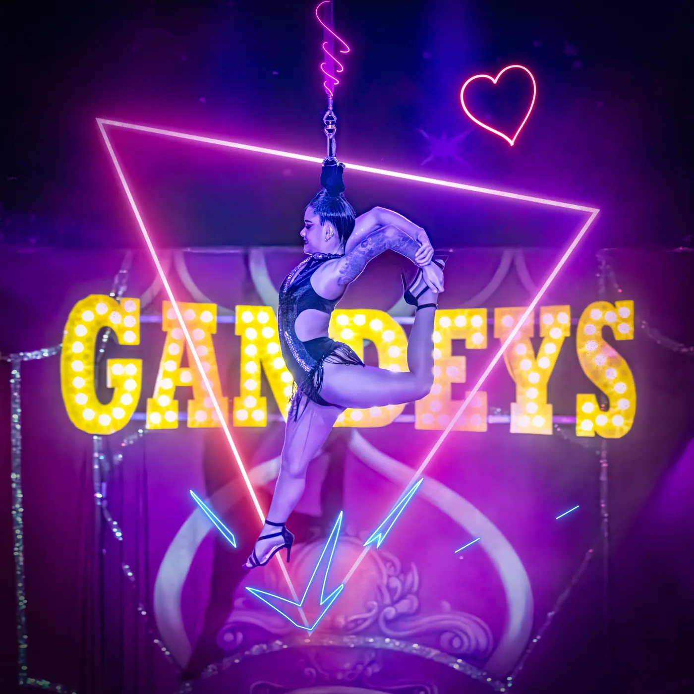 An aerialist hanging by her hair from a chain in front of the lit Gandeys sign
