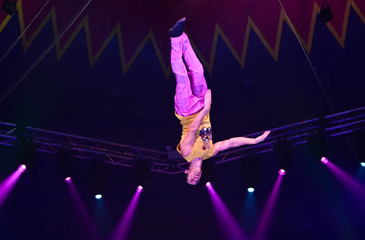 An aerialist in a yellow top flying upside down beneath the Big Top rigging
