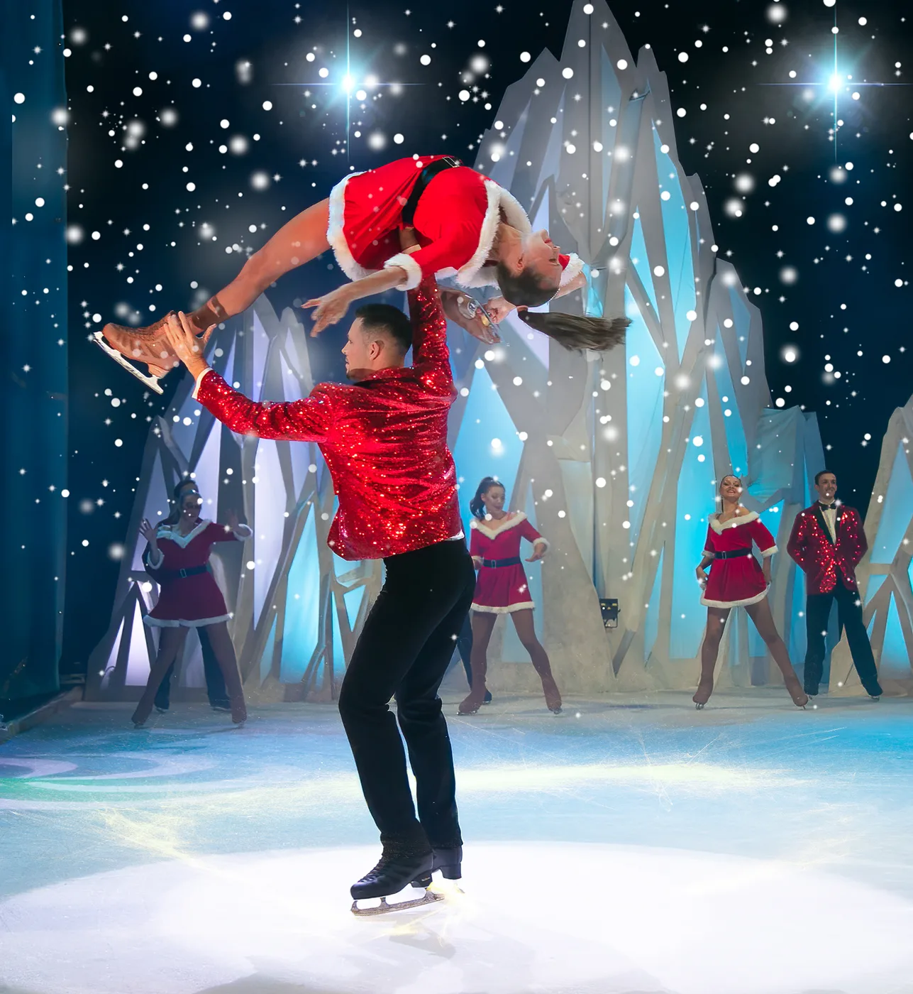 A skater in a red sequinned jacket lifts a partner in a Santa costume above his head, with dancers in red behind